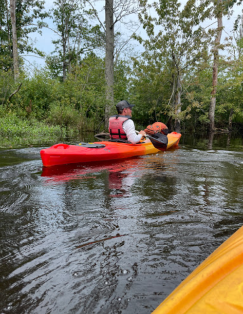 A person in a red and yellow kayak paddles through a calm, tree-lined waterway, wearing a life jacket.