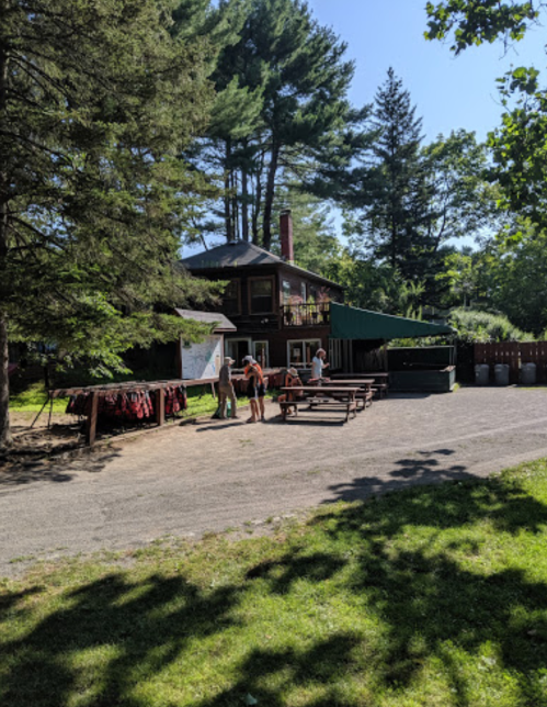 A rustic cabin surrounded by trees, with people gathered outside and picnic tables in the foreground.