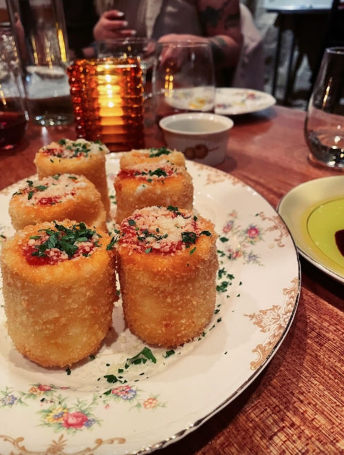 A plate of six golden, crispy croquettes topped with sauce and herbs, set on a decorative floral dish.