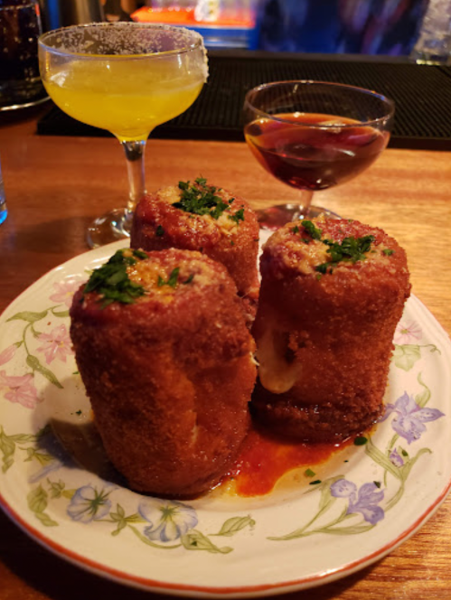 Three crispy, golden-brown croquettes topped with herbs, served on a floral plate with two cocktails in the background.