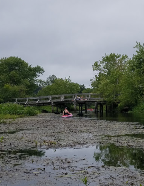 A person in a kayak paddles on a calm, green river under a cloudy sky, with a wooden bridge in the background.