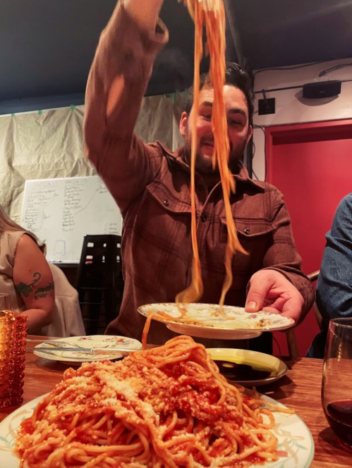 A person in a brown shirt lifts a plate of spaghetti, with a large serving of pasta in front on a wooden table.