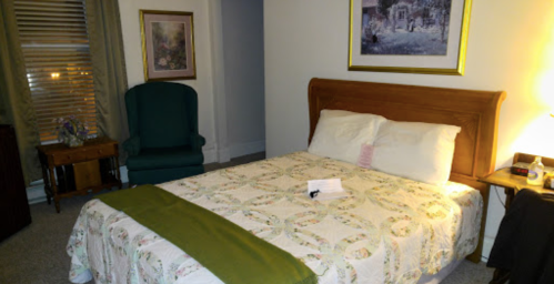 Cozy bedroom with a bed, green throw, armchair, and framed artwork on the walls. Soft lighting creates a warm atmosphere.