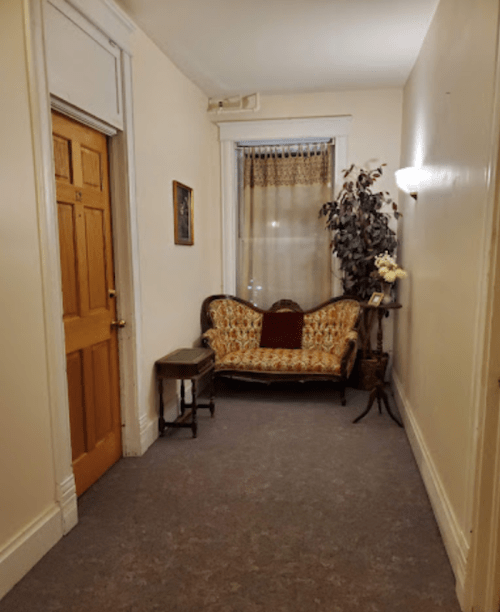 A narrow hallway featuring a vintage floral couch, a small table, and a door, with soft lighting and a window.