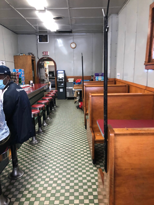 A diner interior with a long counter, red stools, wooden booths, and a checkered floor. A clock is on the wall.