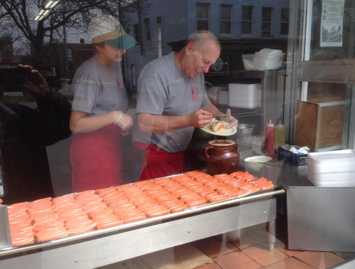 A chef prepares food behind a glass window, with a young assistant and trays of hot dogs in front.