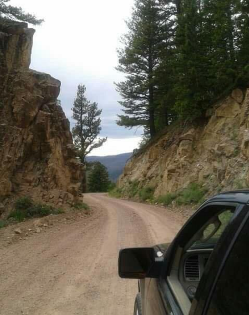 A winding dirt road flanked by rocky cliffs and tall trees, with a vehicle parked at the edge.
