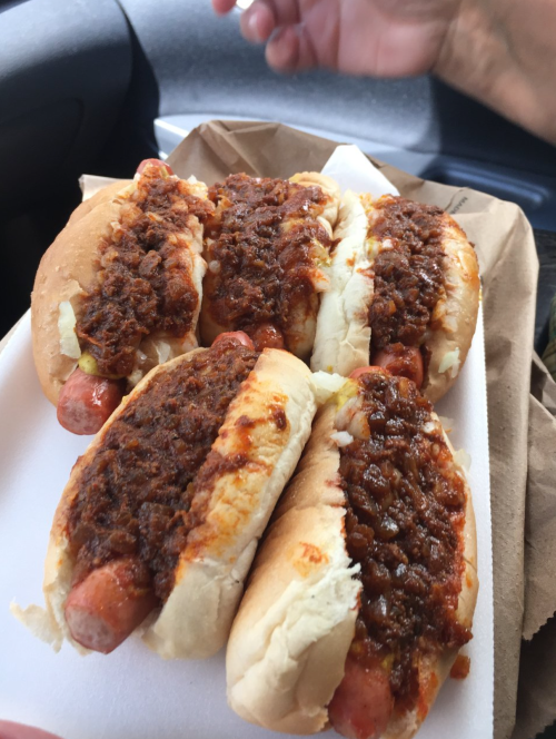 Four hot dogs topped with chili and onions, served in a white tray, with a hand visible in the background.