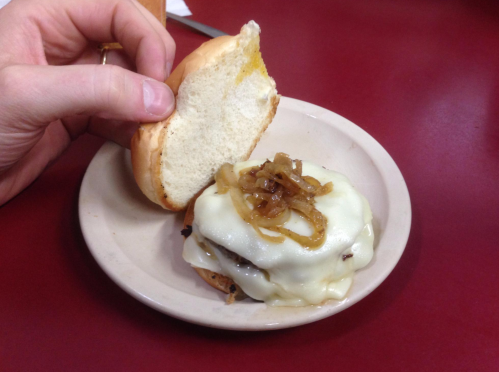 A hand holds a bun above a plate with a cheeseburger topped with melted cheese and sautéed onions.
