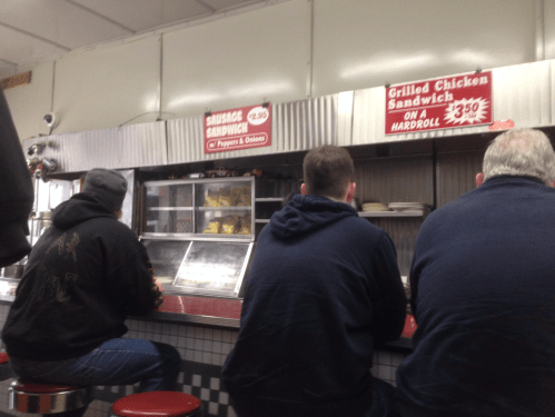 Three customers sit at a counter in a diner, looking at a menu with sandwich options displayed above.