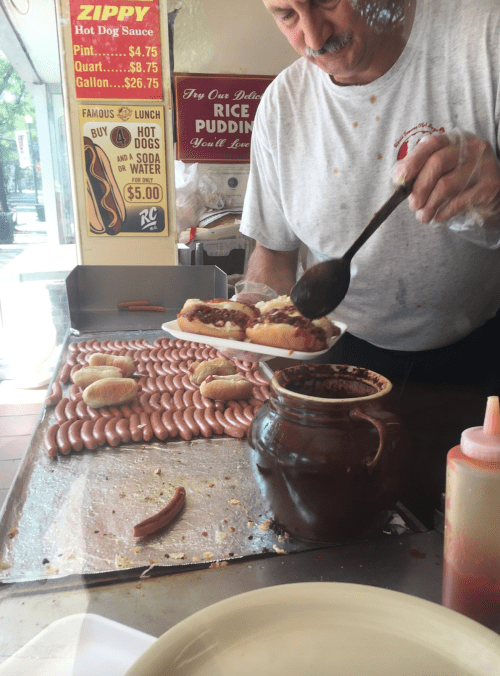 A vendor serves hot dogs topped with sauce from a large pot, with more hot dogs visible in the background.