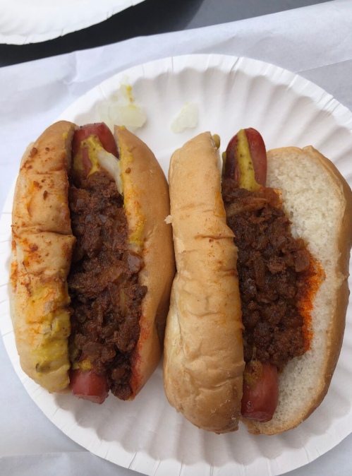 Two hot dogs in buns topped with chili and mustard, served on a paper plate.