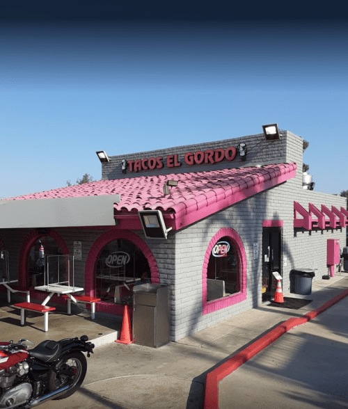 Exterior of Tacos El Gordo, a colorful restaurant with a pink roof and open sign, featuring outdoor seating and a motorcycle nearby.