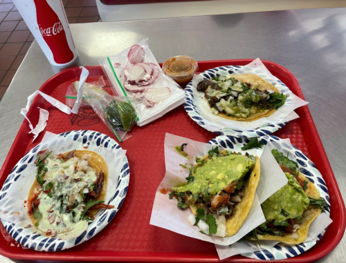 A red tray with four tacos topped with various ingredients, a side of radishes, lime, and a small container of sauce.