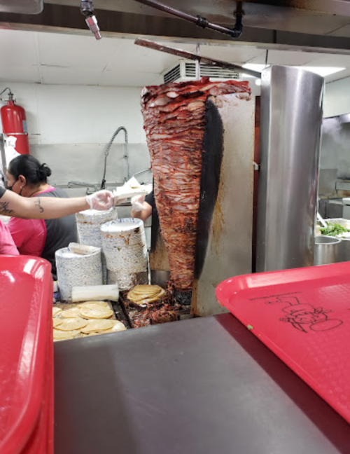 A vertical spit of marinated meat cooking in a kitchen, with tortillas and toppings ready for serving.