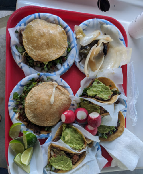 A red tray with several tacos, garnished with lettuce and avocado, alongside lime wedges and radishes.