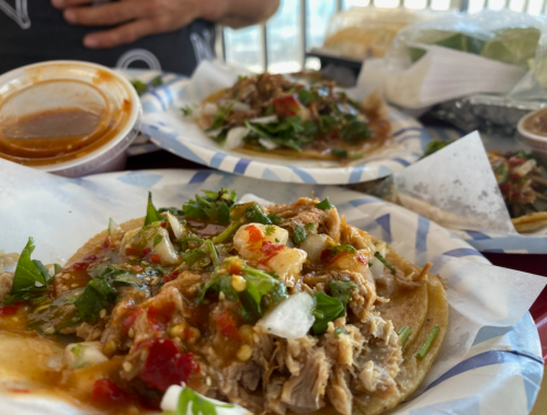 Two plates of tacos topped with meat, onions, cilantro, and salsa, with a side of sauce in a cup.