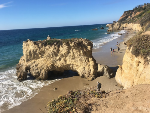 A scenic beach with rocky cliffs, gentle waves, and people walking along the shore under a clear blue sky.