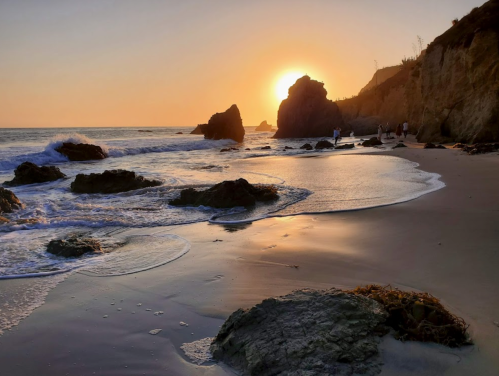 A serene beach at sunset, with gentle waves, rocky formations, and a warm golden glow on the horizon.