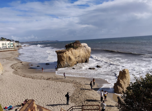 A sandy beach with people walking near a large rock formation and gentle waves under a cloudy sky.
