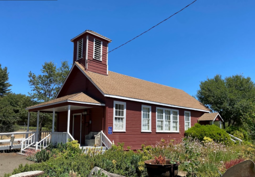 A red, one-story building with a bell tower, surrounded by greenery and blue sky.