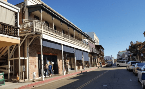 Historic buildings line a sunny street, with people walking and parked cars along the curb.