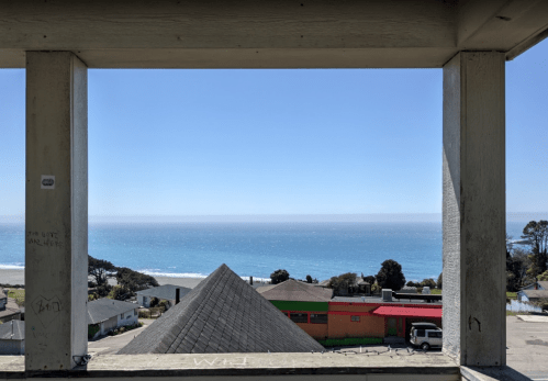 View of the ocean and coastline framed by a concrete structure, with a clear blue sky and distant hills.
