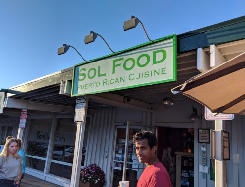 A restaurant sign reads "Sol Food - Puerto Rican Cuisine" with people nearby in a casual outdoor setting.