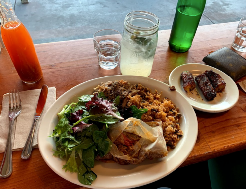 A plate with salad, rice, and a wrapped dish, alongside a drink and a side of grilled food on a wooden table.
