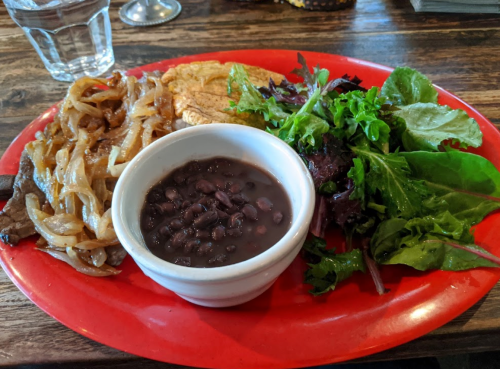 A red plate with grilled meat, sautéed onions, black beans in a bowl, and a side of mixed greens.