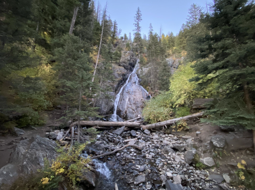 A serene waterfall cascades down rocky terrain, surrounded by lush green trees and autumn foliage.