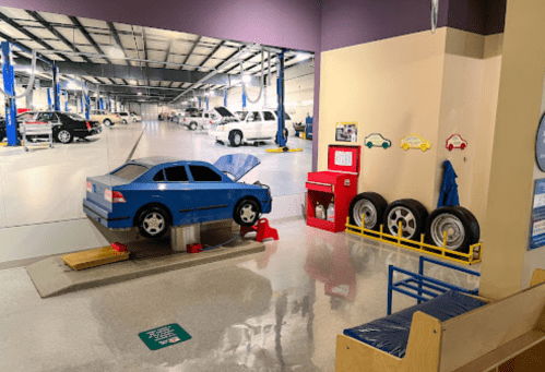 A blue toy car with an open hood in a play area, surrounded by tires and a backdrop of a car repair shop.