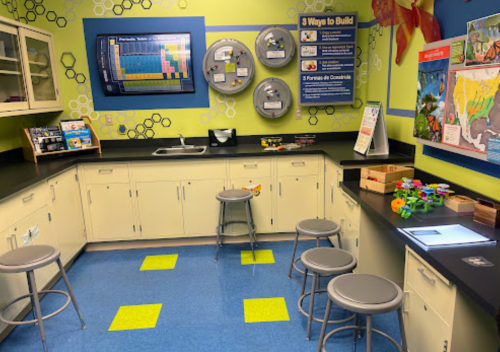 A colorful classroom with a sink, stools, and educational posters on the walls, featuring a hands-on learning area.