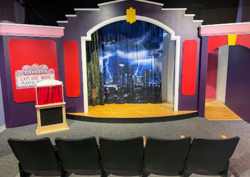 A colorful theater stage with a backdrop of a stormy city skyline, featuring empty seats in front.