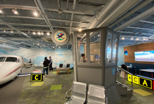 A modern aviation exhibit featuring a control tower model, seating area, and a small aircraft in a spacious room.