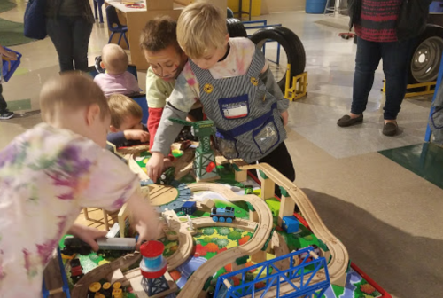Children play with a colorful train set, exploring tracks and toys in a lively indoor environment.