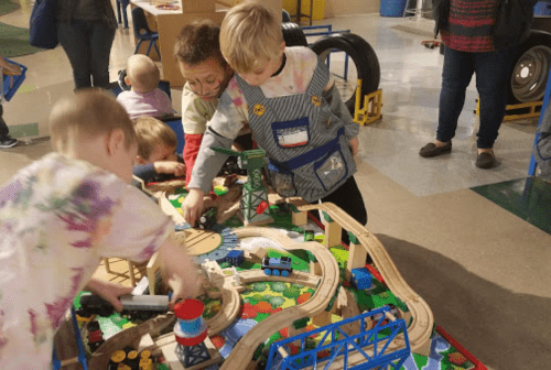 Children play with a colorful train set, exploring tracks and toys in a lively indoor environment.