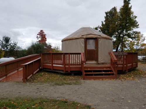 A tan yurt on a wooden deck surrounded by trees, with autumn leaves scattered on the ground.