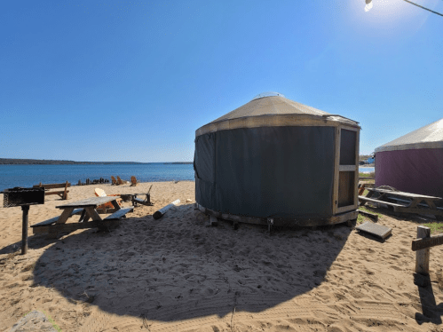 A yurt on a sandy beach by a lake, with picnic tables and chairs in the background under a clear blue sky.