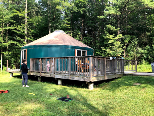 A green yurt on a wooden deck surrounded by trees, with a person standing nearby on the grass.