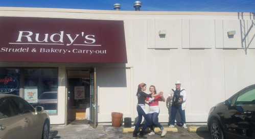 Two women dance outside Rudy's Strudel & Bakery while a man plays an accordion nearby. Sunny day, storefront visible.