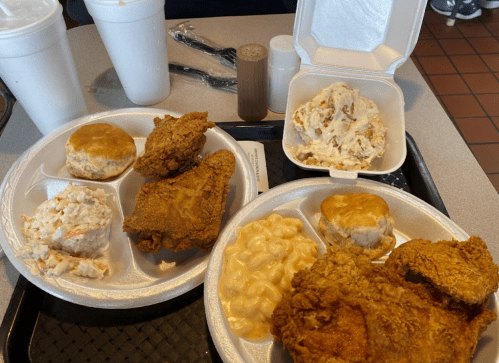 Two trays of fried chicken with sides of coleslaw, macaroni and cheese, biscuits, and drinks in styrofoam cups.