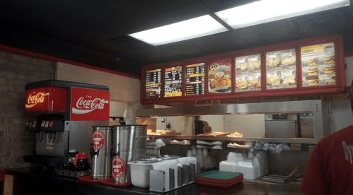 Interior of a fast-food restaurant featuring a menu board, drink dispenser, and food preparation area.