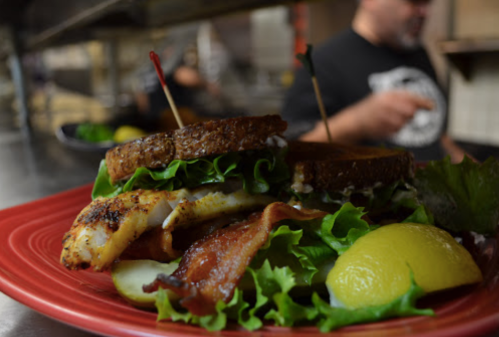 A close-up of a sandwich with grilled fish, bacon, lettuce, and lemon on a red plate, with a kitchen background.