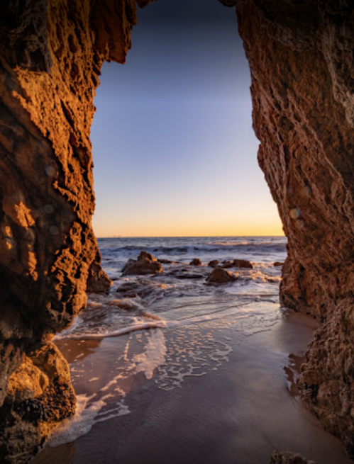 A serene beach view through rocky cliffs at sunset, with gentle waves lapping at the shore.