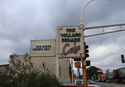Sign for "The Village Square Cafe" with a message about hiring summer help, set against a cloudy sky.