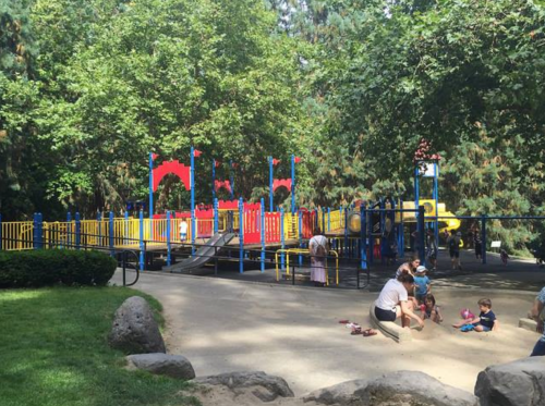 A colorful playground with slides and climbing structures, surrounded by trees, with children playing and adults supervising.