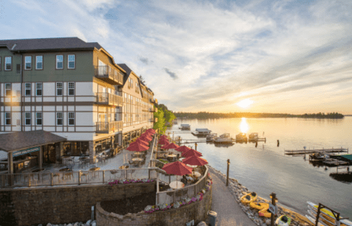 Sunset over a calm lake, with a waterfront hotel, kayaks, and outdoor seating under red umbrellas.