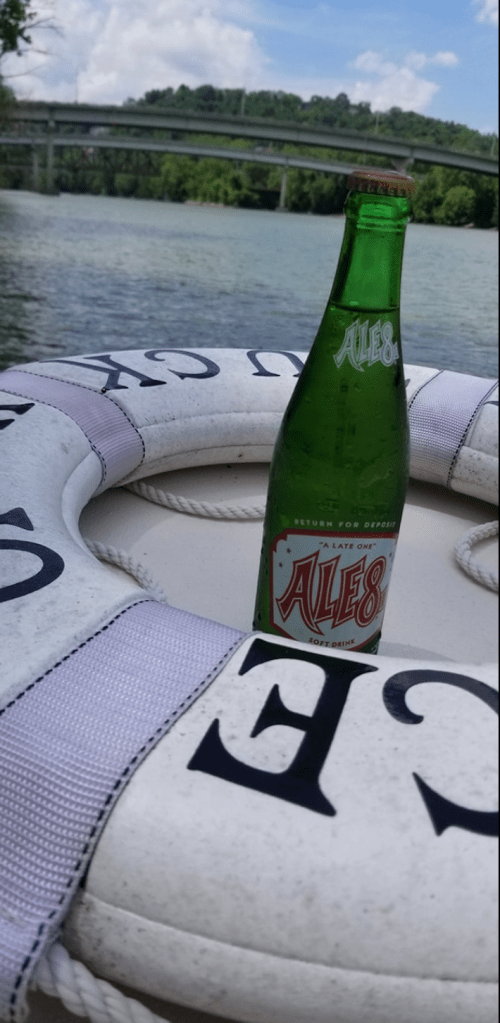 A bottle of Ale-8 soda rests on a life preserver near a river, with a bridge and trees in the background.