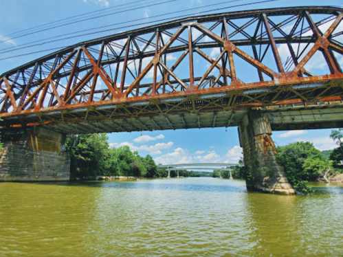 Rusty metal bridge arching over a river, surrounded by lush greenery and a clear blue sky.